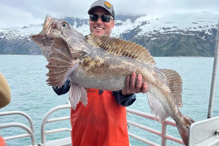 Person in orange overalls holding a large fish on a boat with snowy mountains in the background.