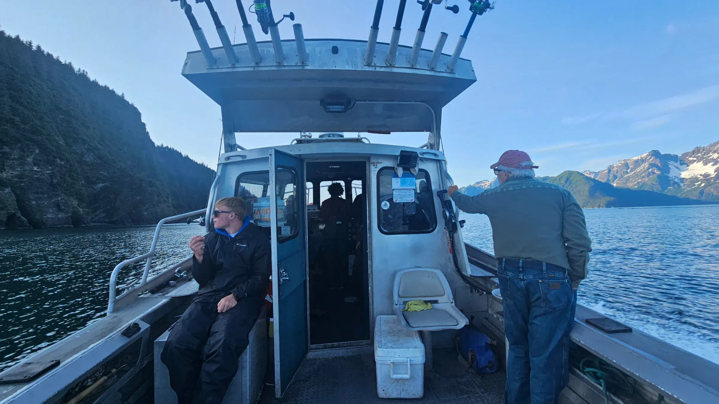 Two people on a boat with fishing rods, surrounded by mountain scenery.