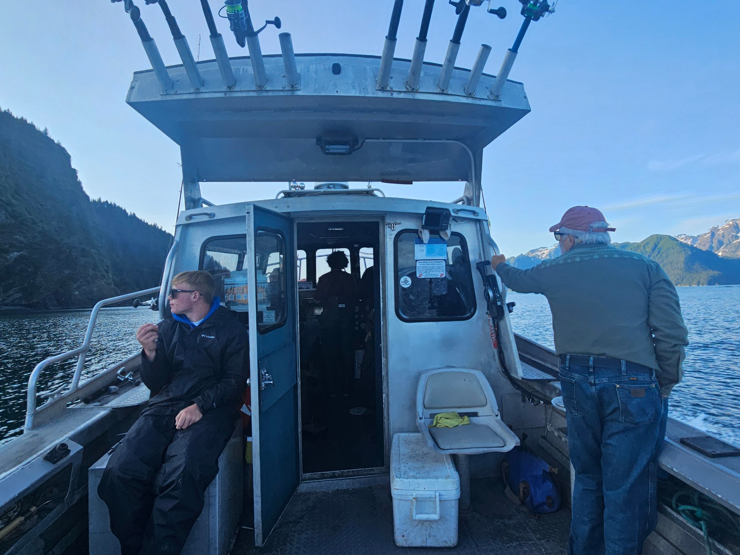 Two people on a boat with fishing rods, surrounded by mountain scenery.