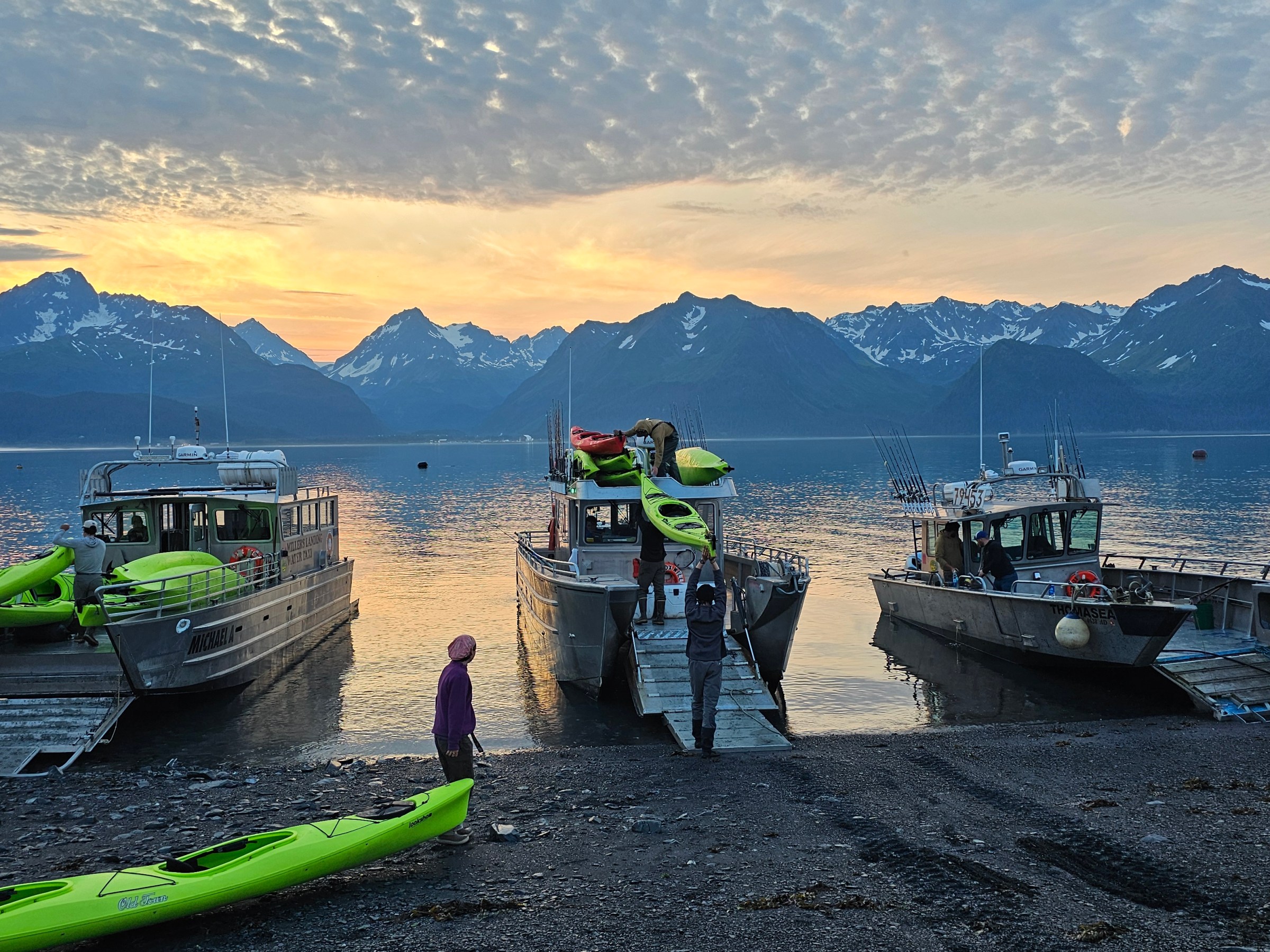Three boats docked with kayaks near a lake and snowy mountains at sunset, people on shore.