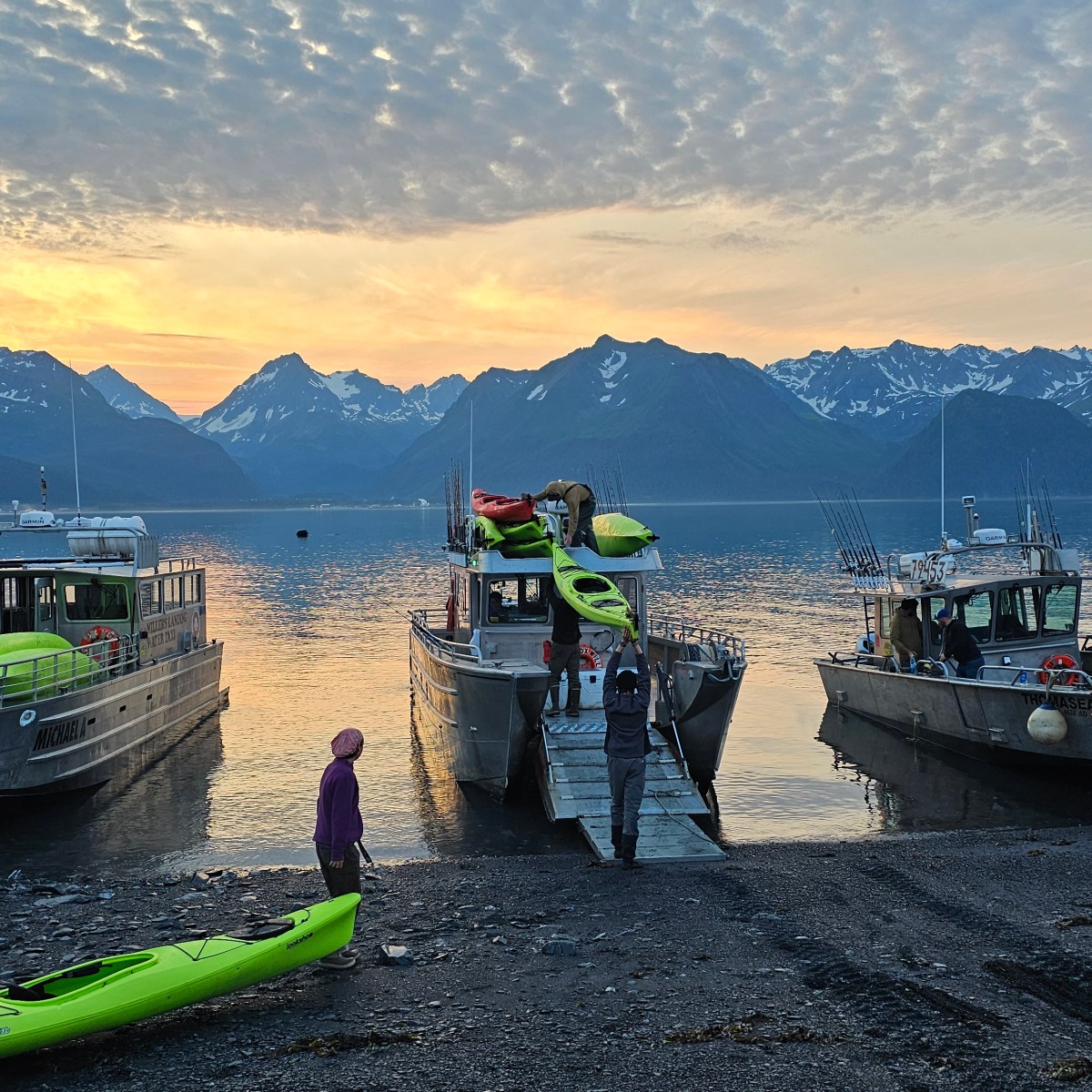 Three boats docked with kayaks near a lake and snowy mountains at sunset, people on shore.