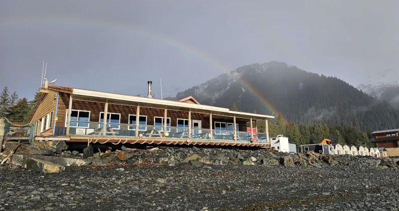 Mountain lodge with a rainbow over a rocky landscape and forested hills.