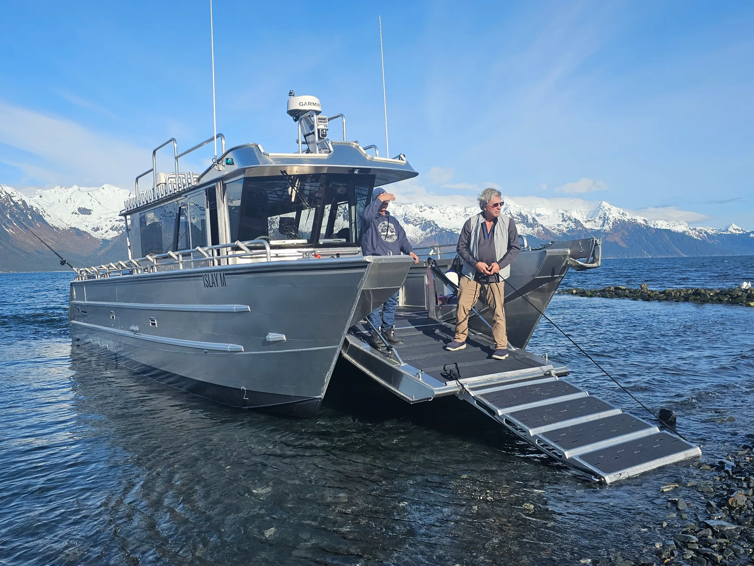 Two people stand on a boat ramp by the shore with snowy mountains in the background.