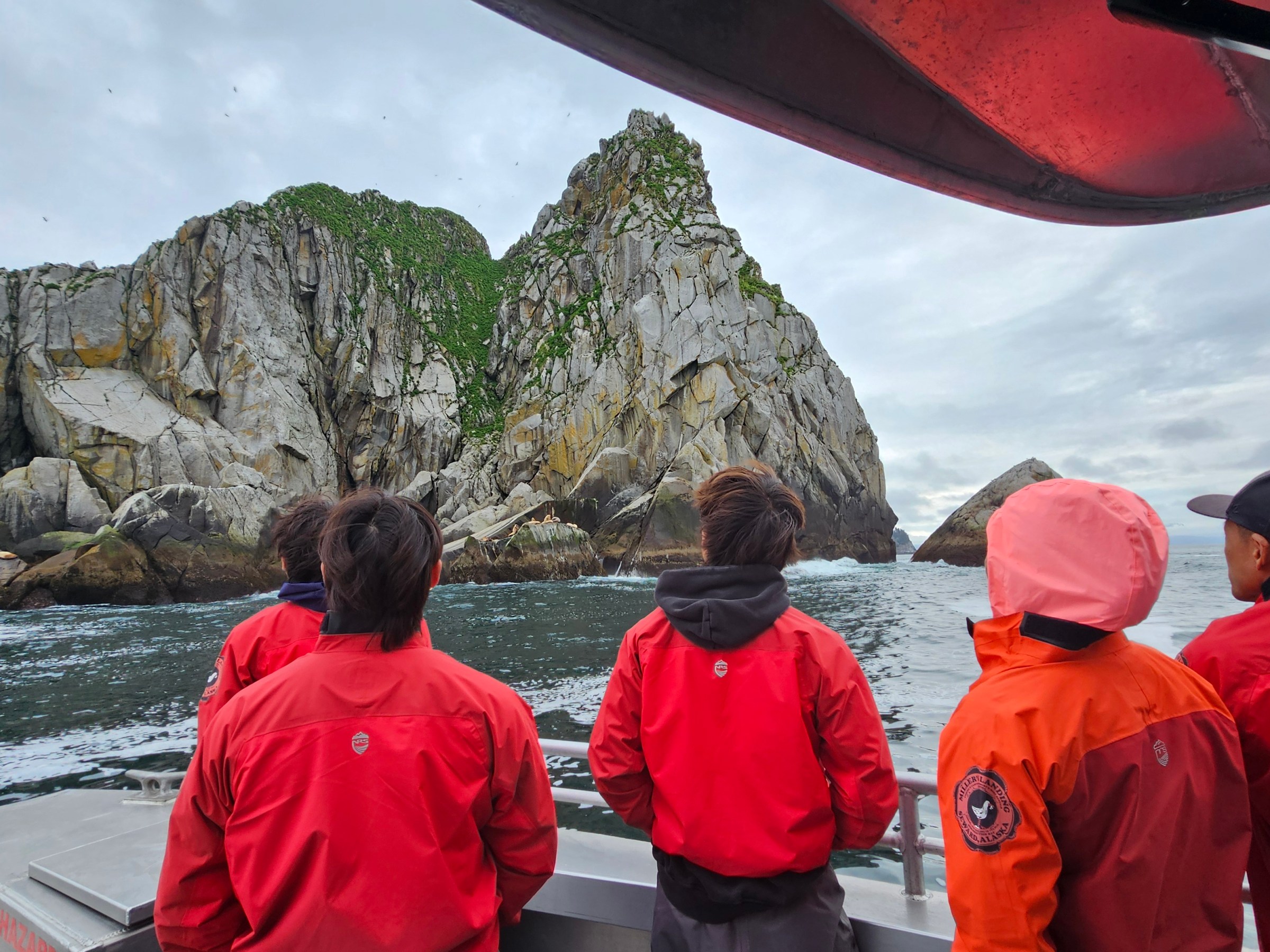 People in red jackets on a boat, looking at a large rocky island with greenery.