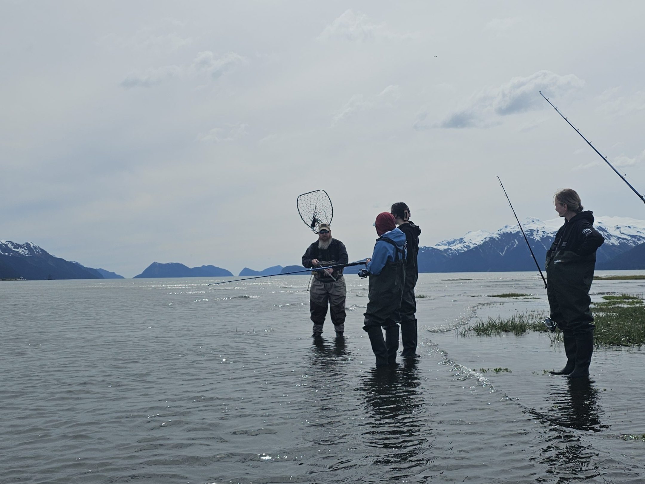 Four people fishing in shallow water, surrounded by mountains under a cloudy sky.