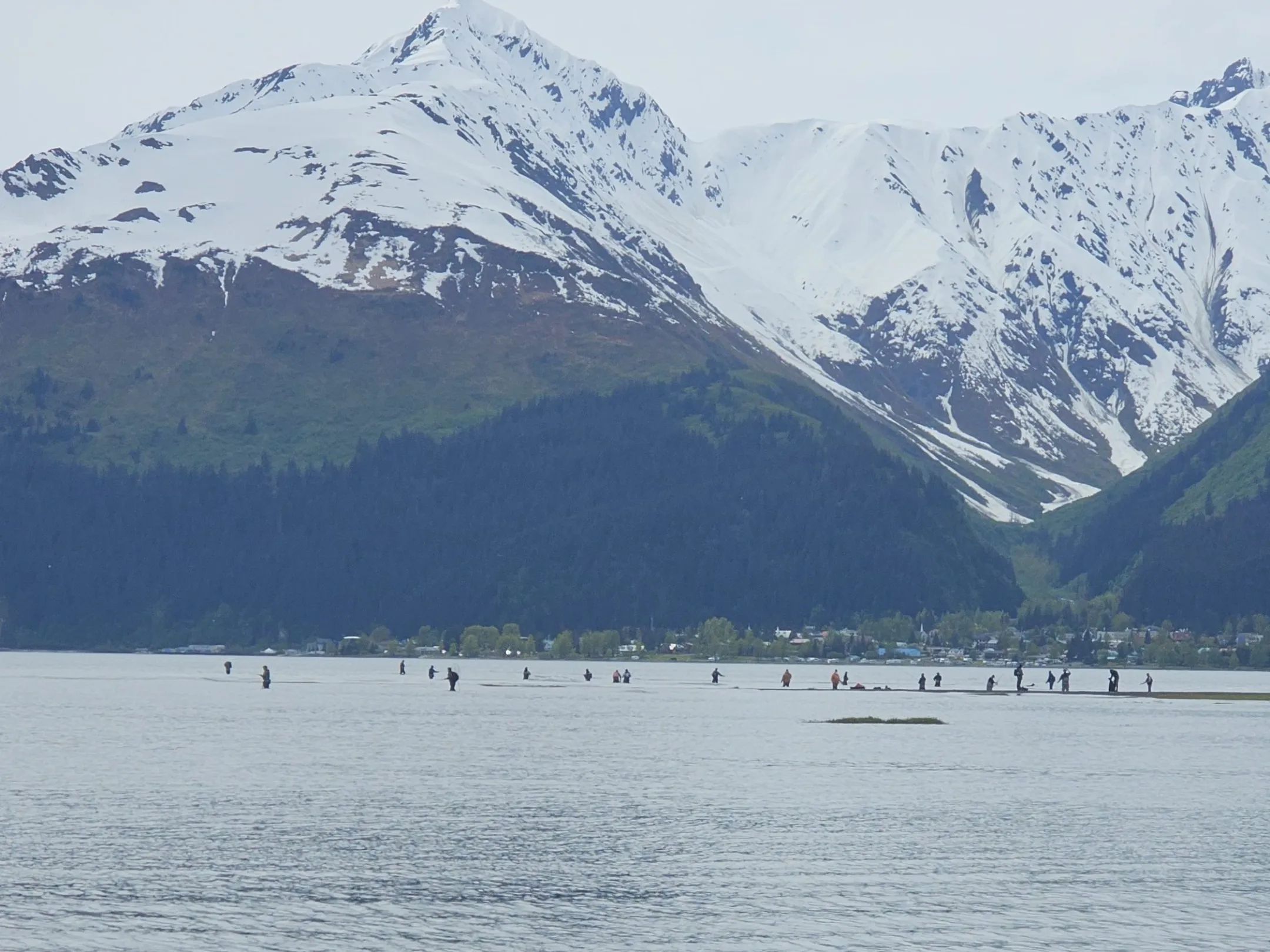Snow-capped mountains with a lake and people fishing in the foreground.