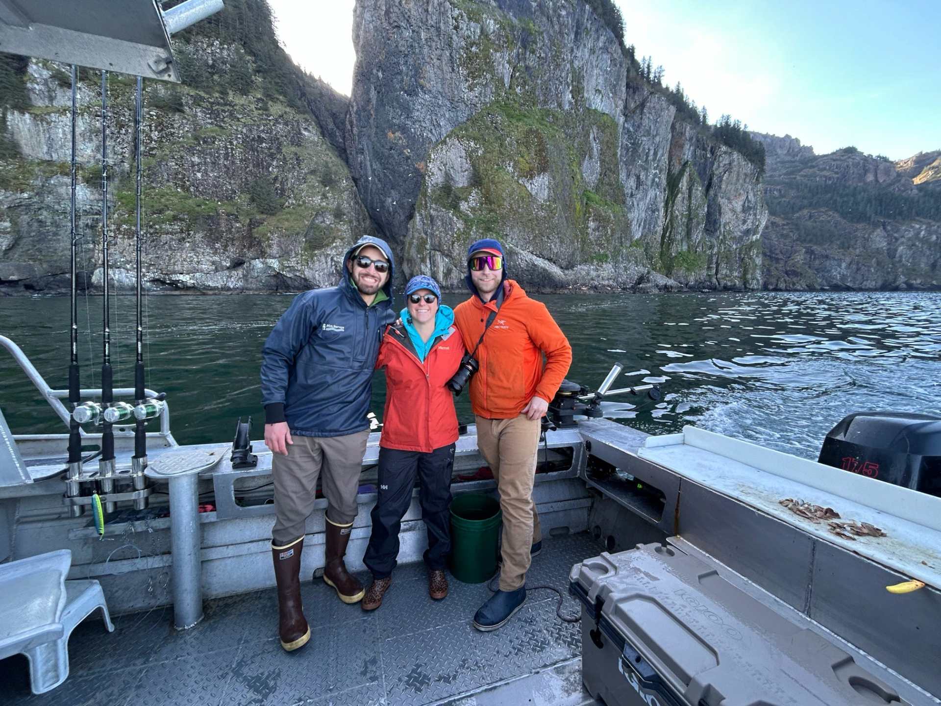 Three people posing on a boat with fishing rods, rocky cliffs in the background.