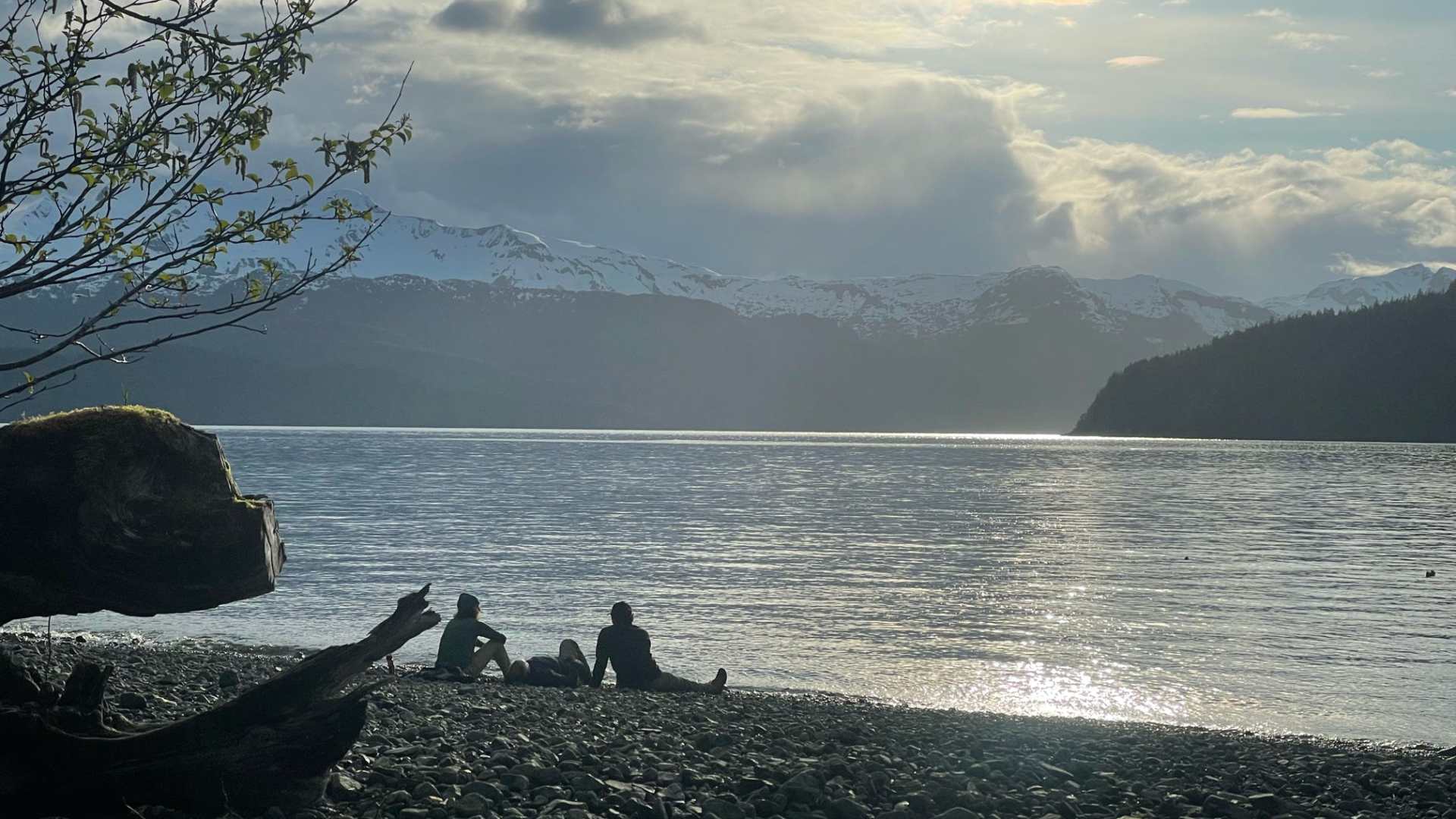 Two people sitting on a rocky beach by a lake with mountains and cloudy sky in the background.