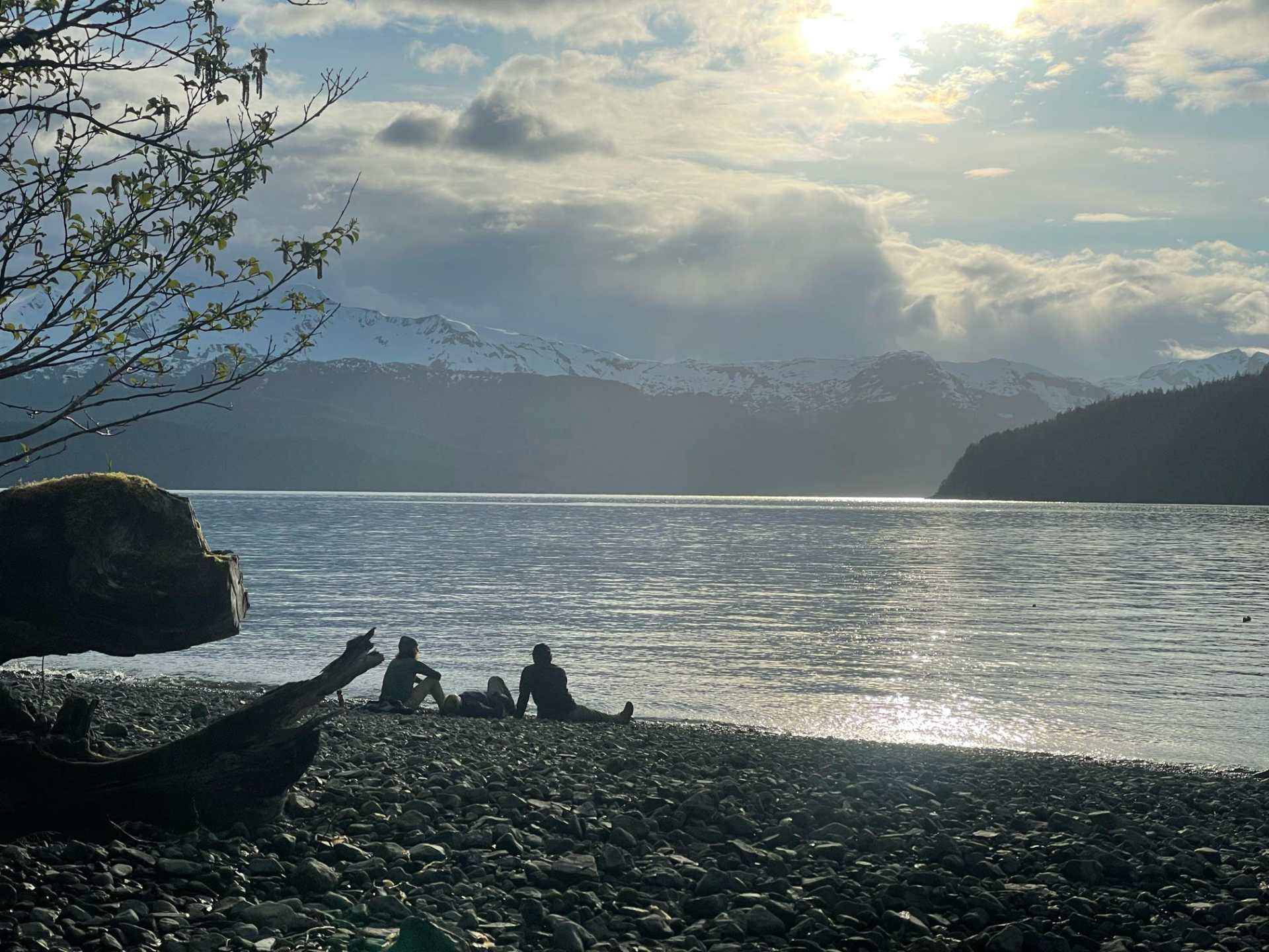 Two people sitting on a rocky beach by a lake with mountains and cloudy sky in the background.