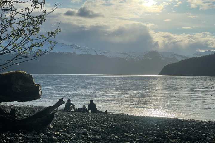 Two people sitting on a rocky beach by a lake with mountains and cloudy sky in the background.