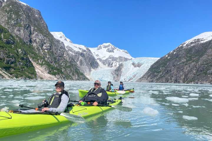 People kayaking on a glacial lake surrounded by mountains and icebergs on a sunny day.