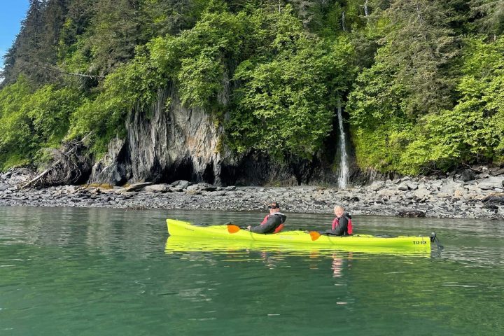 Two people kayaking near a forested cliff with a small waterfall.