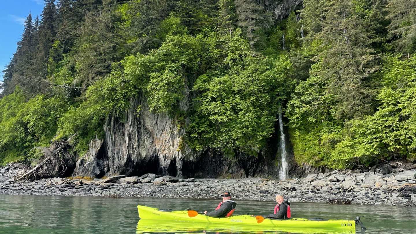 Two people kayaking near a forested cliff with a small waterfall.