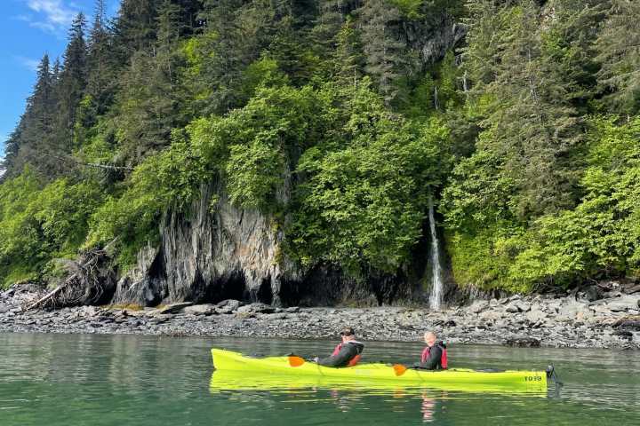 Two people kayaking near a forested cliff with a small waterfall.