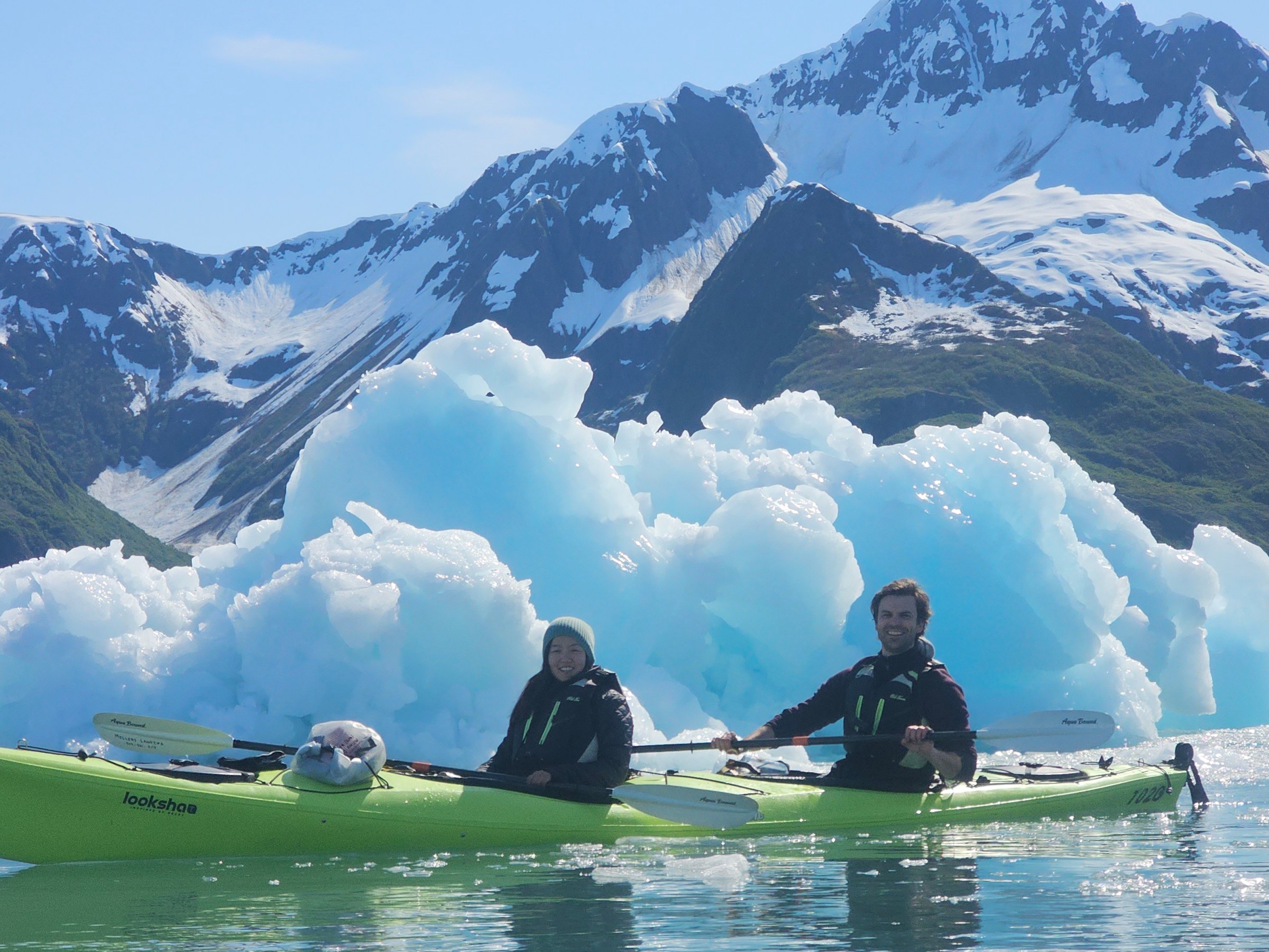 Two people kayaking near a large iceberg with snowy mountains in the background.