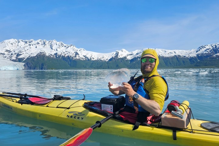 Person kayaking on a calm glacier lake, holding up ice chunk, wearing yellow gear, snow-capped mountains in background.