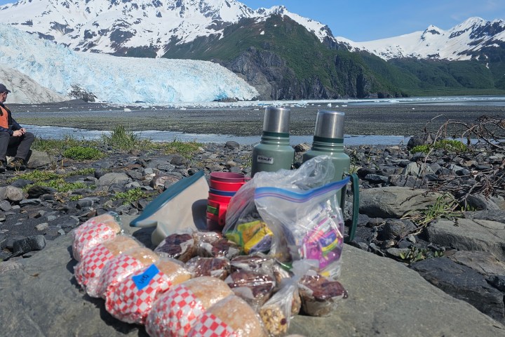 Person sitting by a glacier with snacks, thermoses, and mountains in the background.