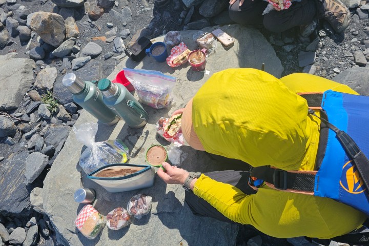 Person in yellow jacket having a picnic on rocks with thermoses and food containers.