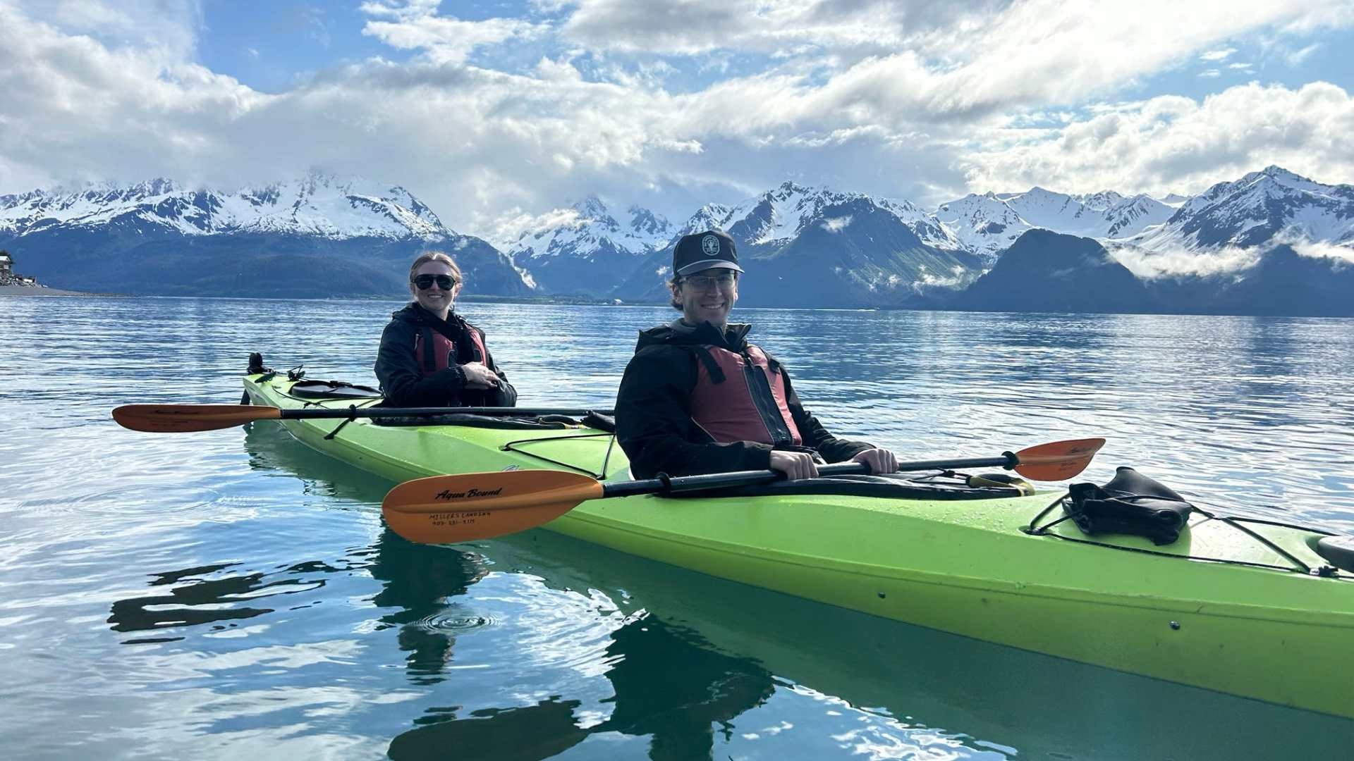 Two people kayaking on calm water with snow-capped mountains in the background.