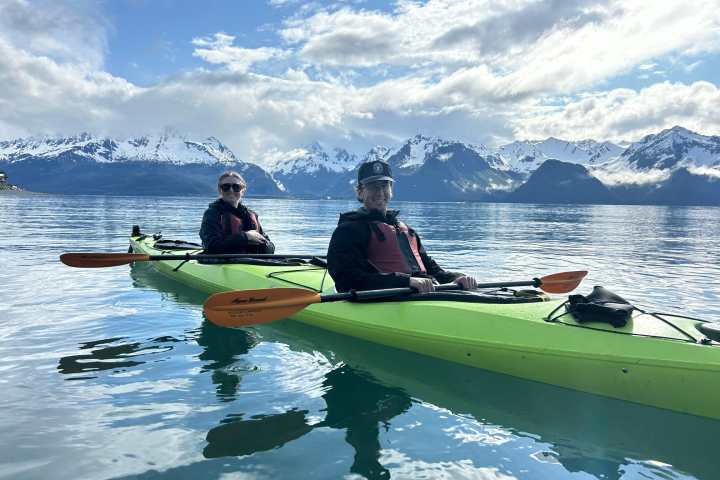 Two people kayaking on calm water with snow-capped mountains in the background.