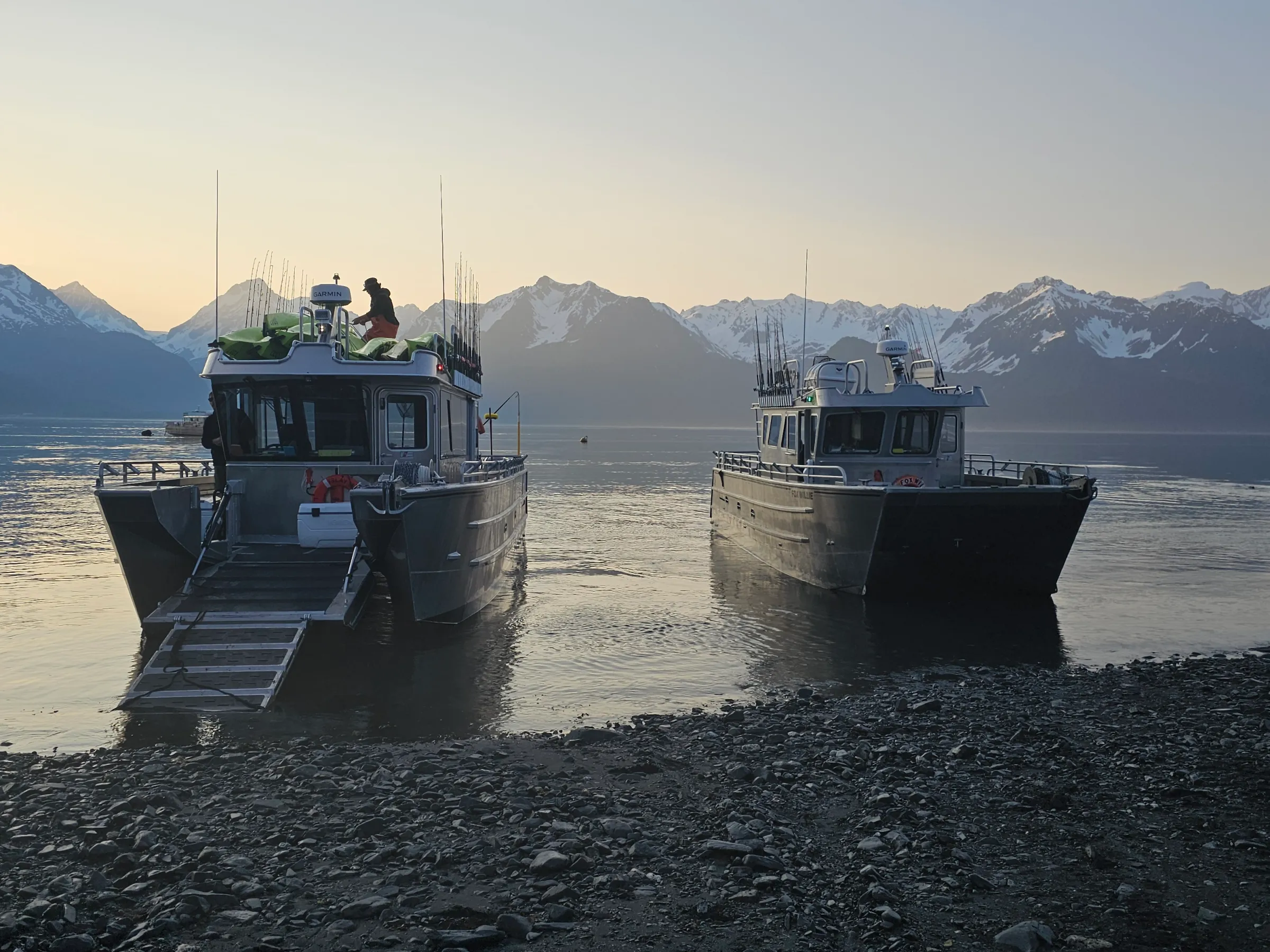 Two boats on a pebbled shore with snow-capped mountains in the background at sunrise.