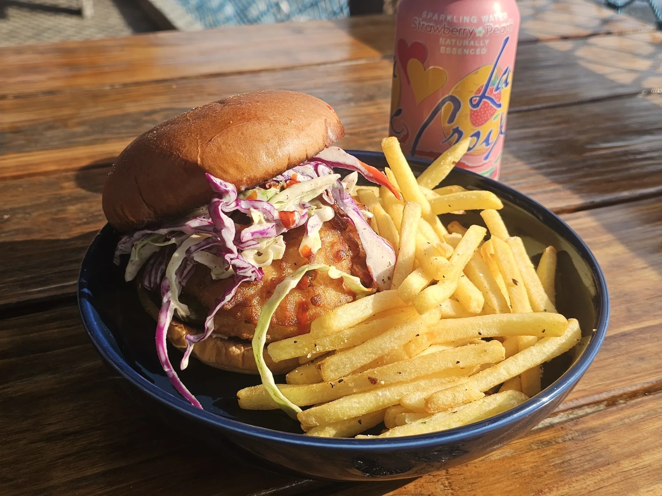 Burger with coleslaw and fries on a plate next to a can of sparkling water on a wooden table outdoors.