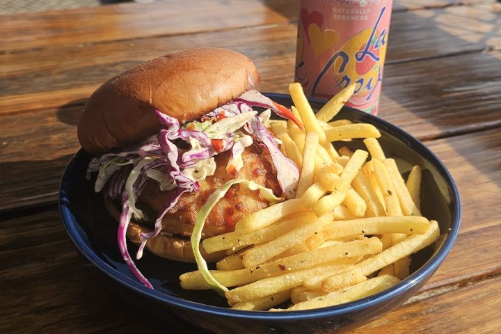 Burger with coleslaw and fries on a plate next to a can of sparkling water on a wooden table outdoors.