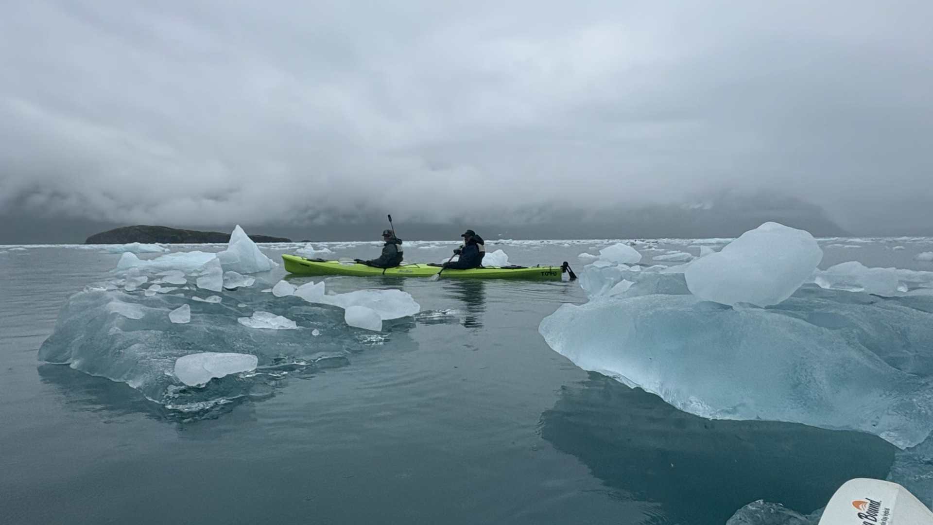 Two people kayaking among icebergs on a calm, cloudy day.