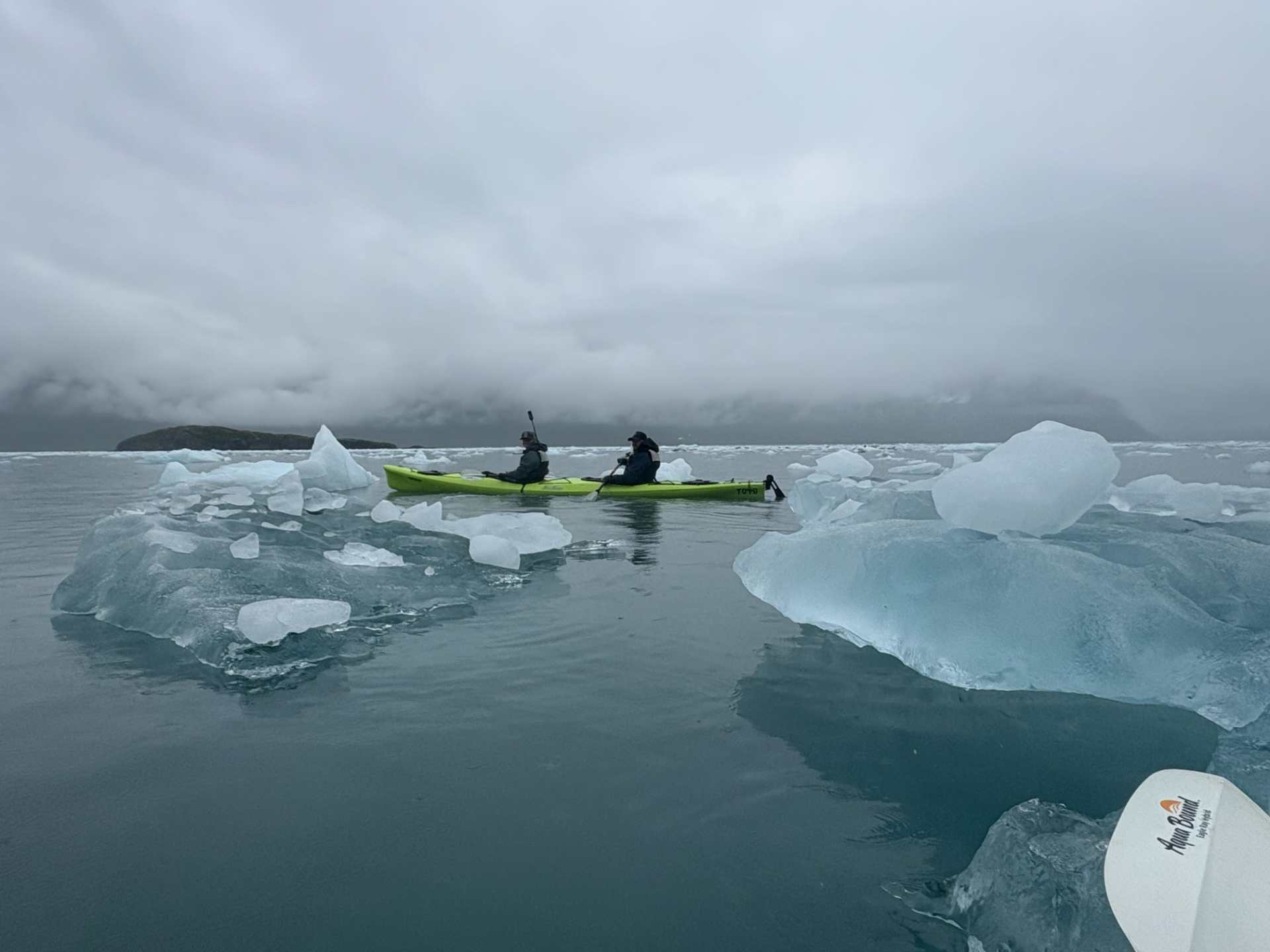 Two people kayaking among icebergs on a calm, cloudy day.