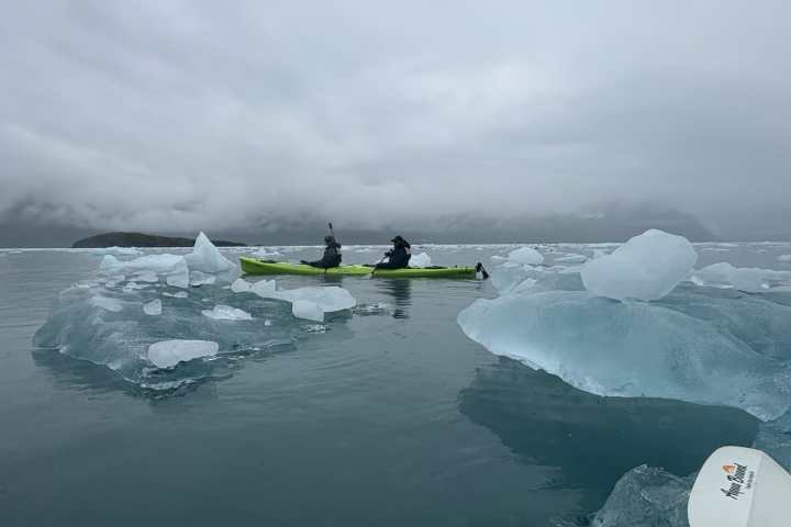 Two people kayaking among icebergs on a calm, cloudy day.