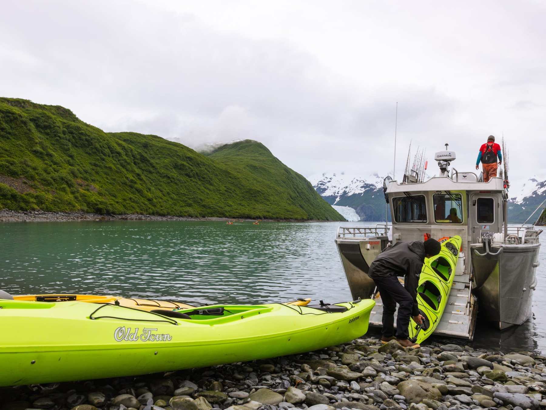 People loading kayaks onto a boat by a rocky shore with green hills in the background.