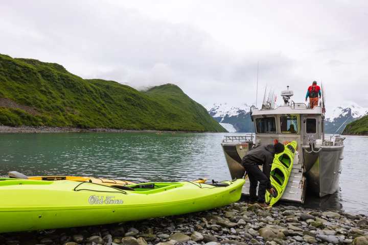 People loading kayaks onto a boat by a rocky shore with green hills in the background.