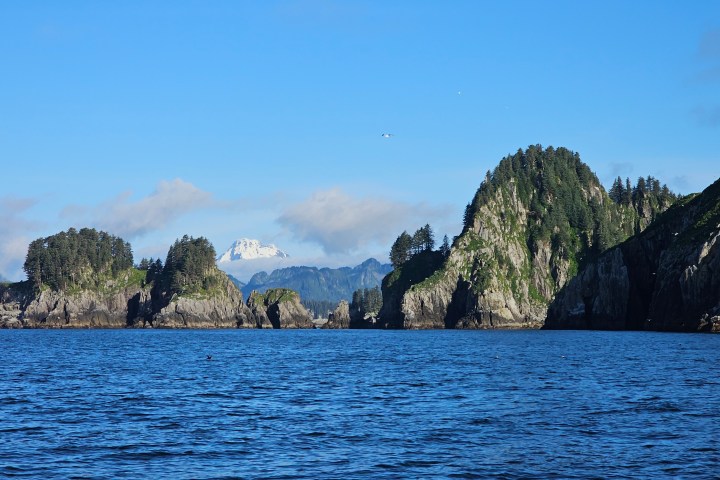 Ocean view of rocky islands with trees, distant snow-capped mountain, and blue sky.