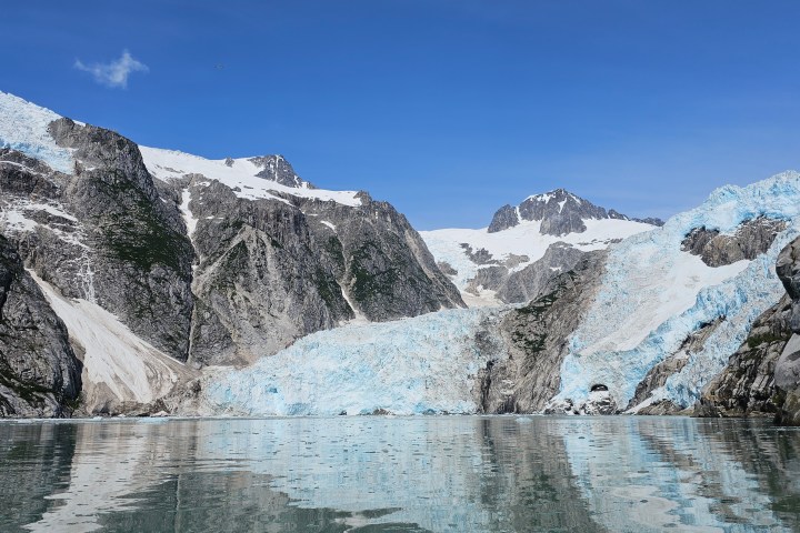 Snow-capped mountain with glacier flowing into a calm fjord under a clear blue sky.