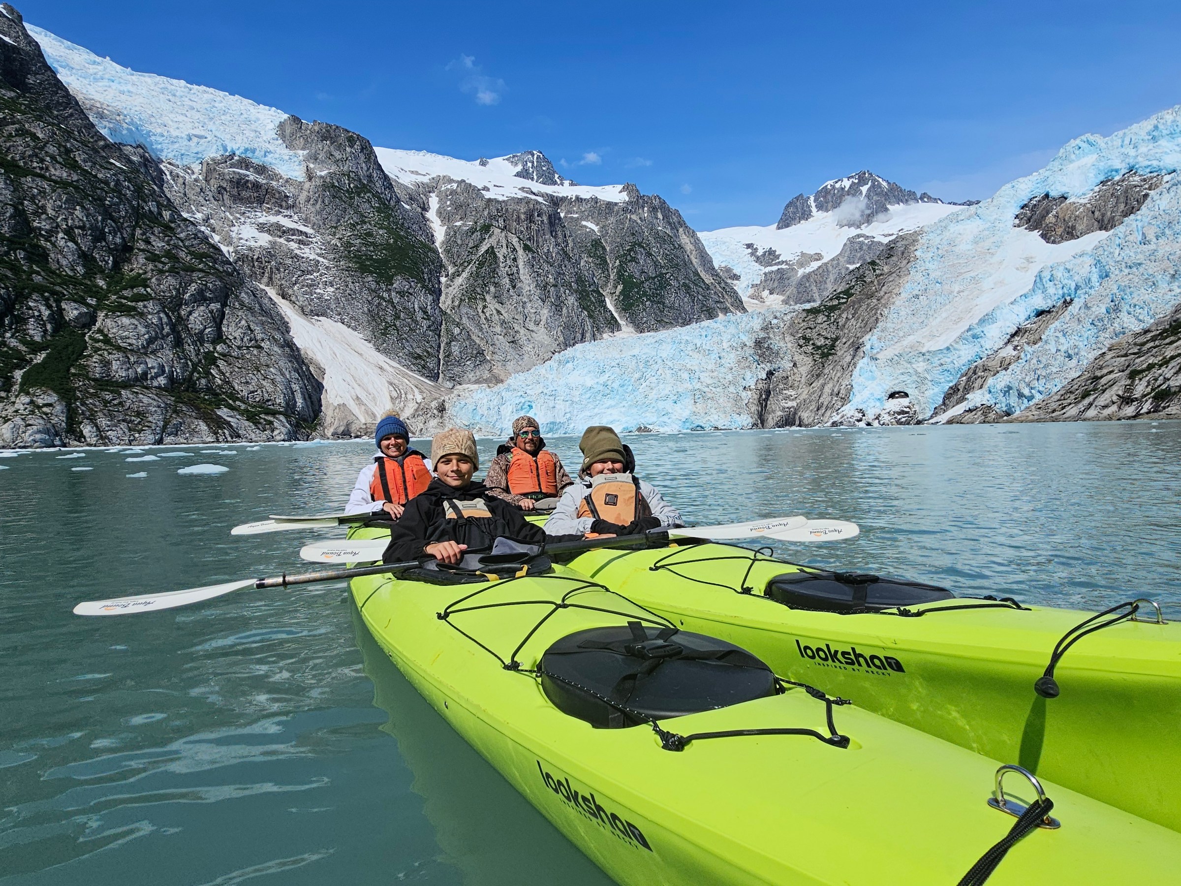 Four people in kayaks on a glacial lake with snowy mountains in the background.