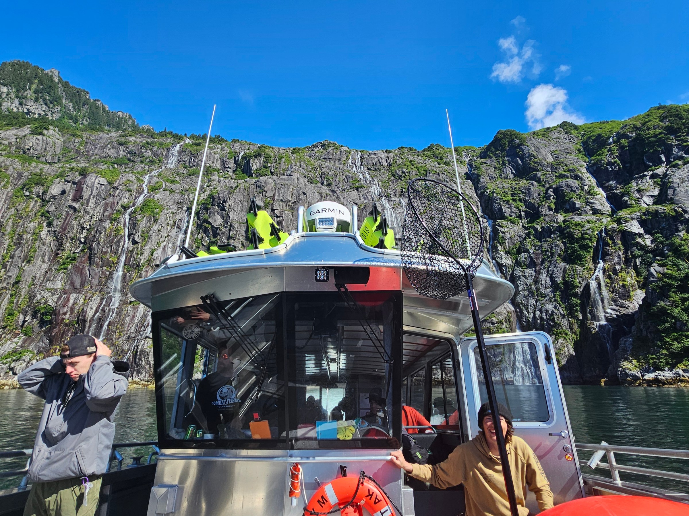 Boat with fishing net in front of rocky cliff and green hills under a blue sky.