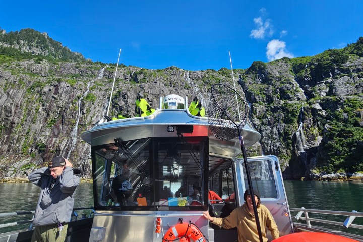 Boat with fishing net in front of rocky cliff and green hills under a blue sky.