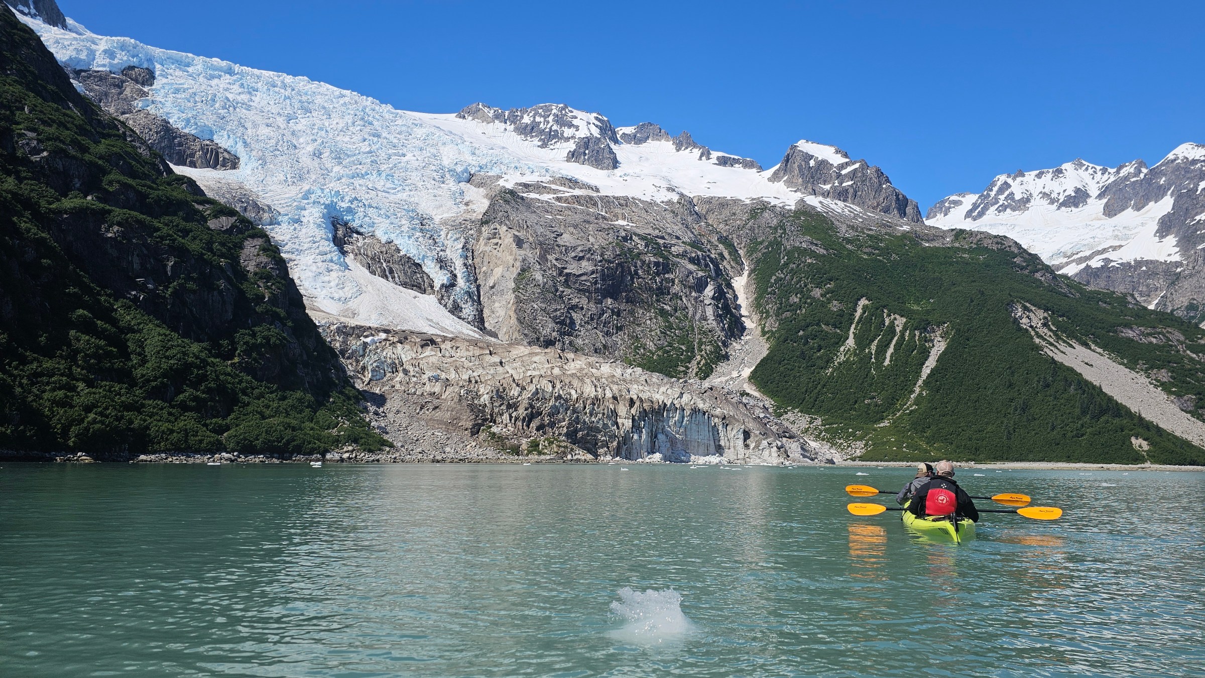 Kayakers on a glacial lake with snow-capped mountains under a clear blue sky.