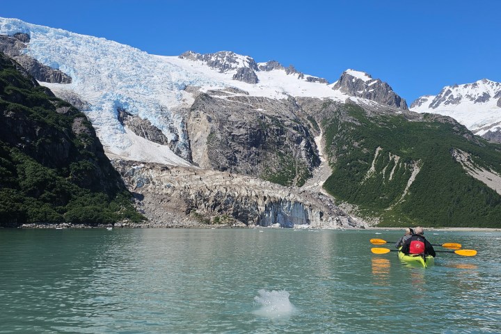 Kayakers on a glacial lake with snow-capped mountains under a clear blue sky.