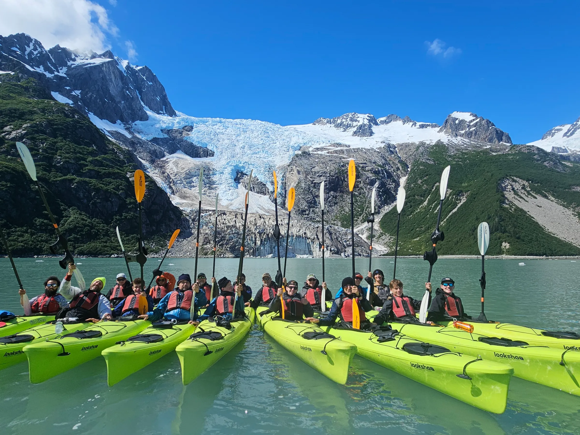 Group of kayakers in yellow kayaks on a lake with a glacier and mountains in the background.