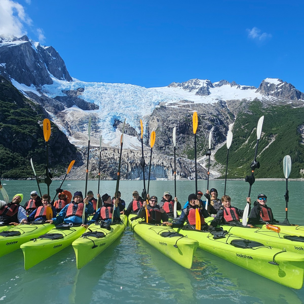 Group of kayakers in yellow kayaks on a lake with a glacier and mountains in the background.