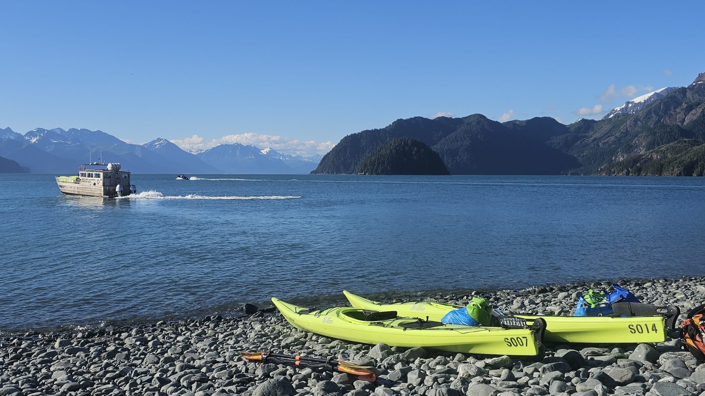 Kayaks and gear on rocky beach, boat on lake, snow-capped mountains in background under clear blue sky.