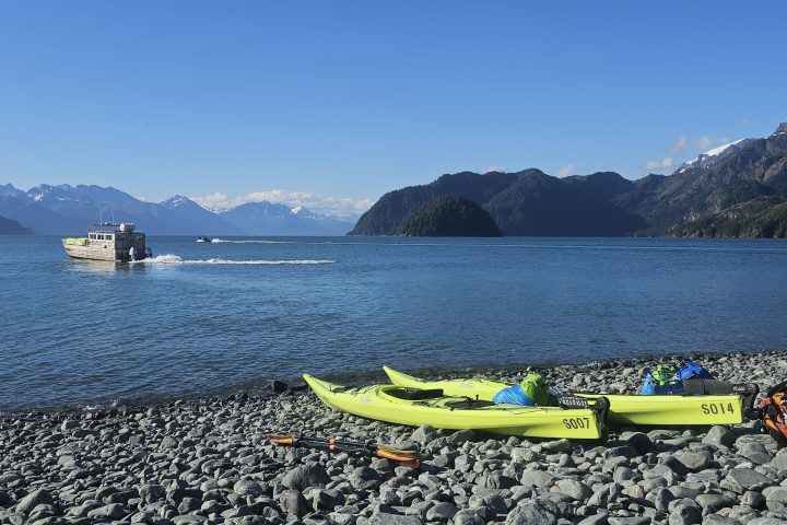Kayaks and gear on rocky beach, boat on lake, snow-capped mountains in background under clear blue sky.