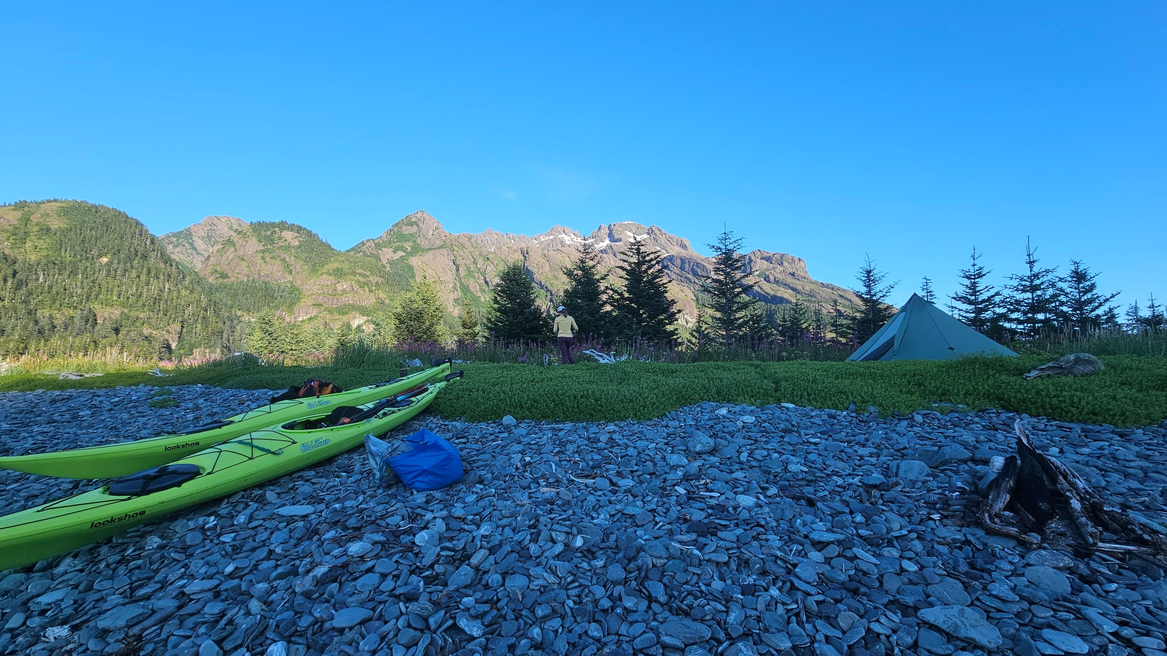 Kayaks and tent on rocky beach with mountains and trees in the background.