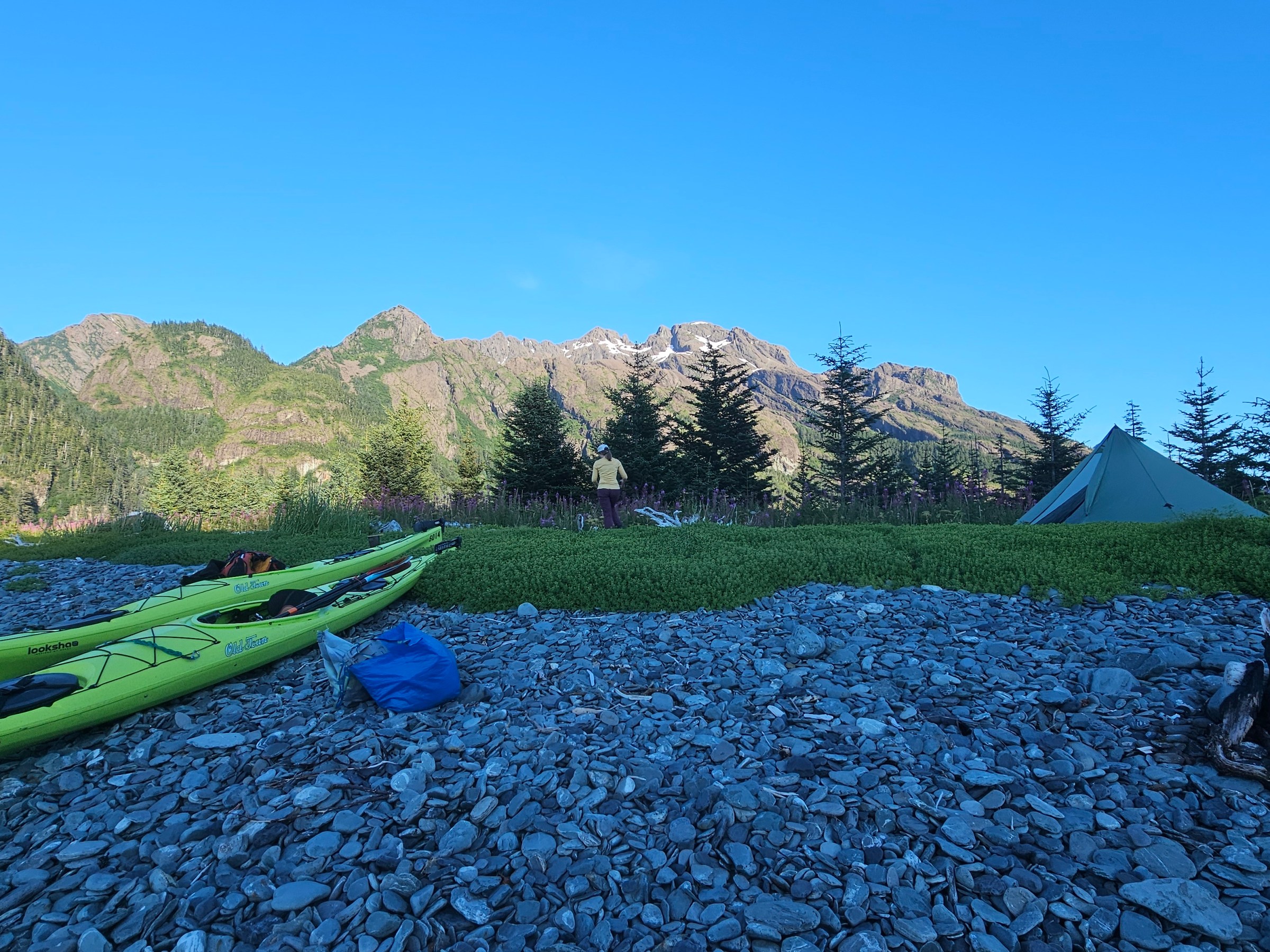Kayaks and tent on rocky beach with mountains and trees in the background.
