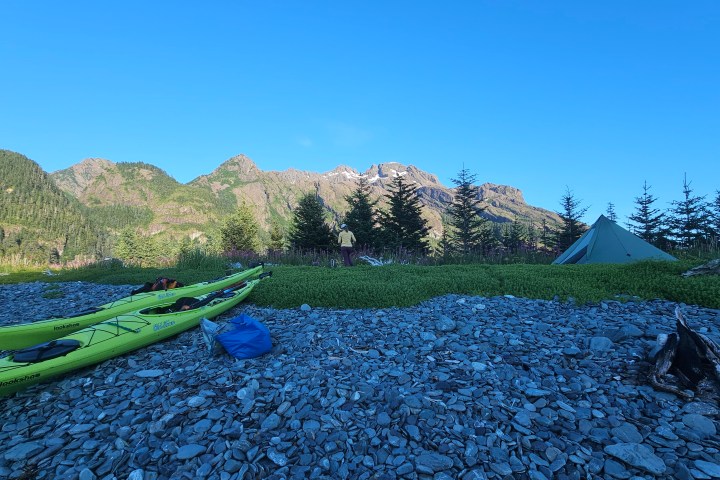 Kayaks and tent on rocky beach with mountains and trees in the background.