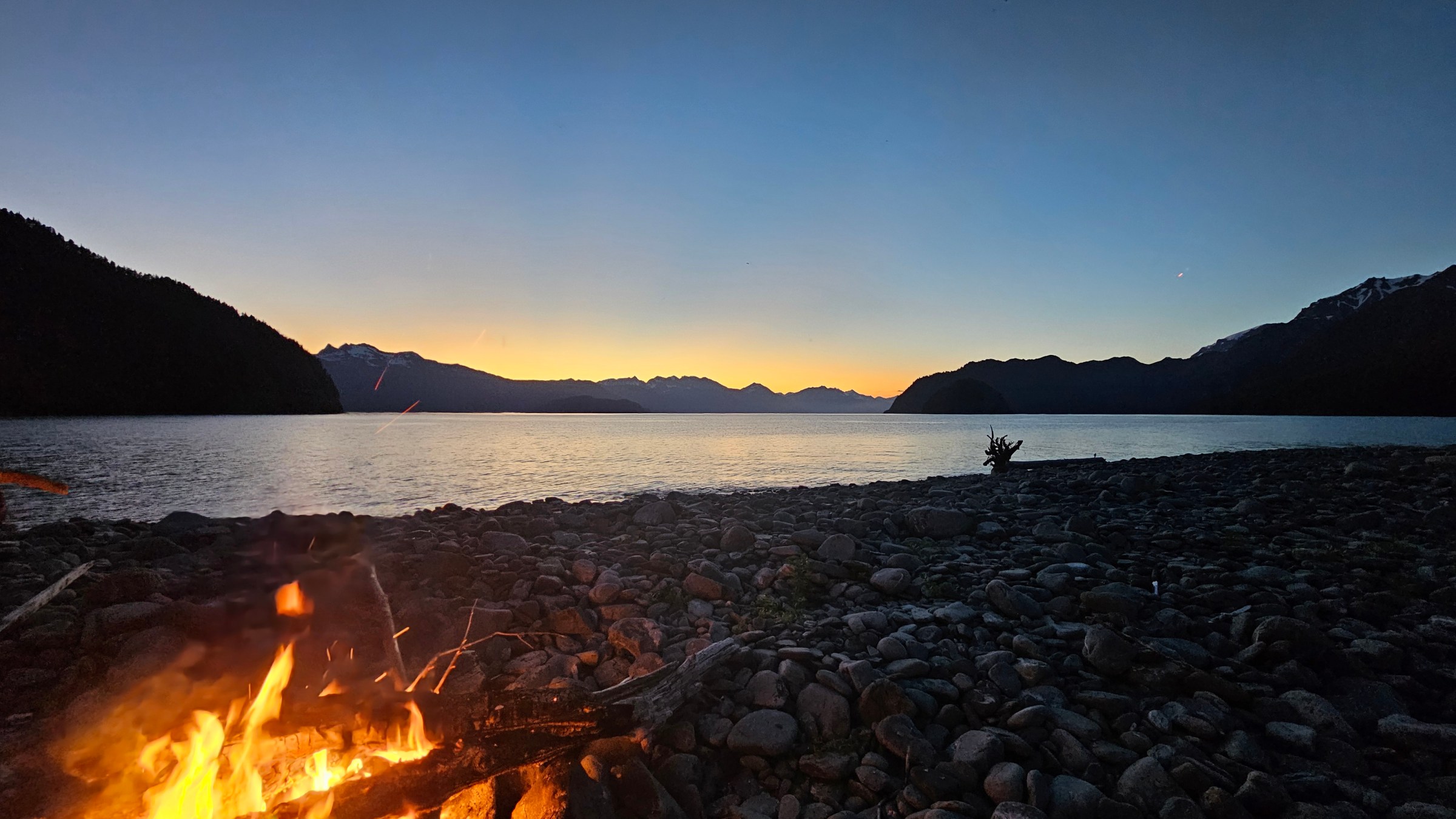 Campfire on rocky lakeshore at dusk with mountains and clear sky.