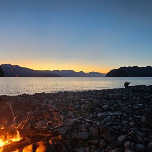 Campfire on rocky lakeshore at dusk with mountains and clear sky.
