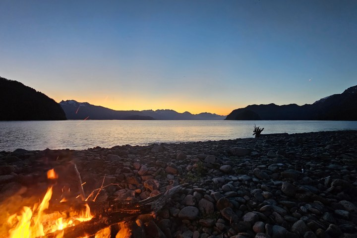 Campfire on rocky lakeshore at dusk with mountains and clear sky.