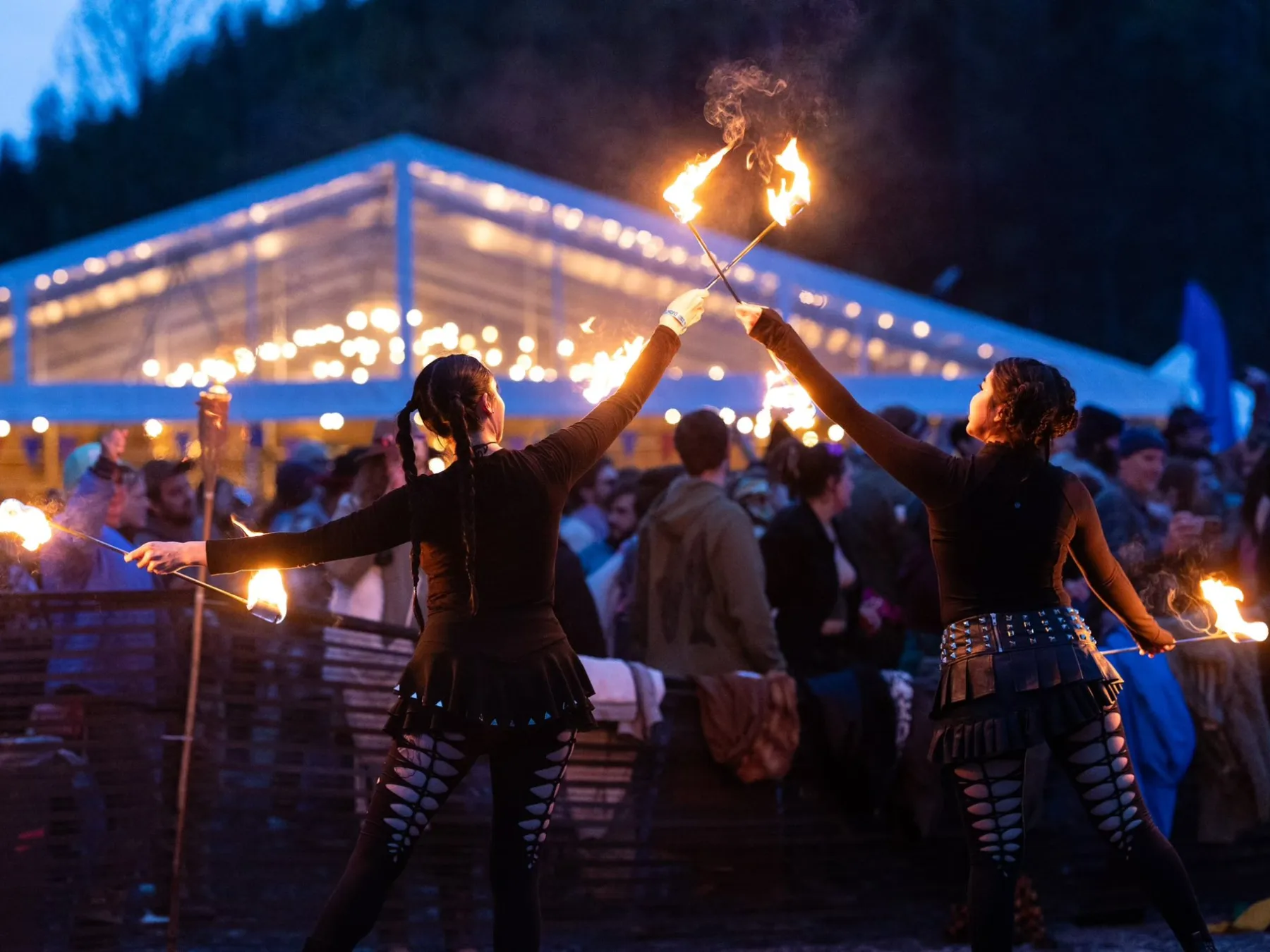 Two performers spinning fire staffs at a night festival, with a lit tent and crowd in the background.